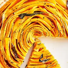 Une magnifique tarte à la courge aux fines lamelles dorées, garnie de feuilles de sauge. Une part a été retirée, révélant la texture appétissante.