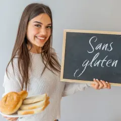 Une jeune femme souriante tient une assiette de pain et un tableau noir "Sans gluten". Elle montre ce qu'il faut manger en cas d'intolérance au gluten.
