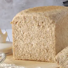 Un pain de farine intégrale, tout juste sorti du four, avec une tranche coupée. Des ingrédients de boulangerie sont disposés autour.