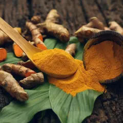 Racines de curcuma et poudre dorée sur une feuille verte, une cuillère en bois débordant de curcuma.