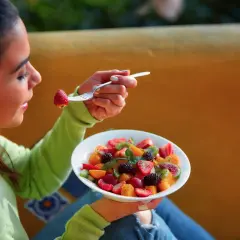 Une jeune femme savoure une salade de fruits colorée, une fraise à la fourchette. La naturopathie suggère quand manger les fruits pour une digestion optimale.