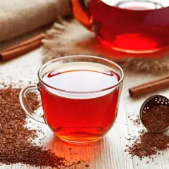 Une tasse de thé rooibos rouge vif, avec des bâtons de cannelle et des feuilles de thé sur une table en bois.