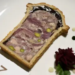 Tranche de pâté en croûte, garnie de pistaches et d'une couche sombre, servie sur une assiette blanche avec des touches de sauce verte et blanche.
