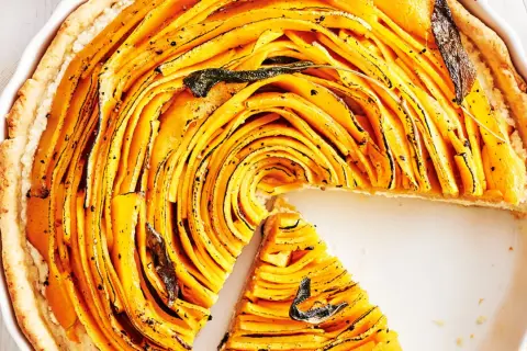 Une magnifique tarte à la courge aux fines lamelles dorées, garnie de feuilles de sauge. Une part a été retirée, révélant la texture appétissante.