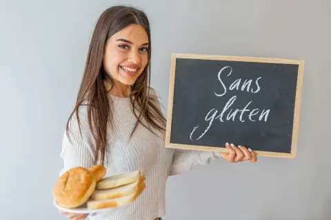 Une jeune femme souriante tient une assiette de pain et un tableau noir "Sans gluten". Elle montre ce qu'il faut manger en cas d'intolérance au gluten.