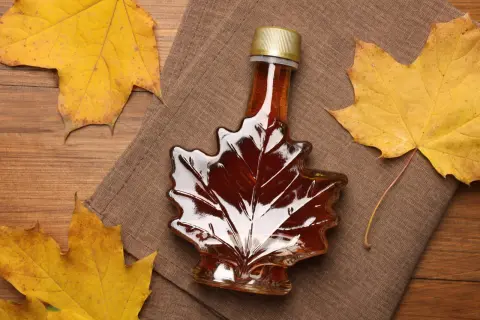 Bouteille en forme de feuille d'érable remplie de sirop d'érable ambré, entourée de feuilles d'automne. Le sirop d'érable, c'est quoi ? Un délice naturel.