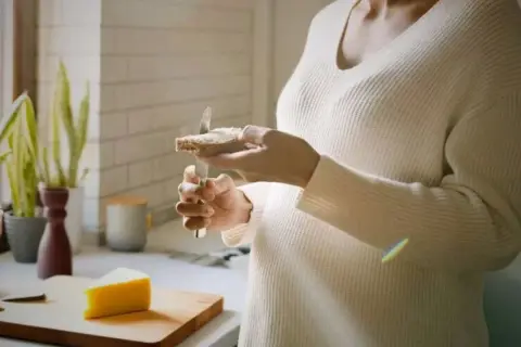 Une personne tartine une tranche de pain avec du fromage au lait cru. Un morceau de fromage est posé sur une planche à découper.