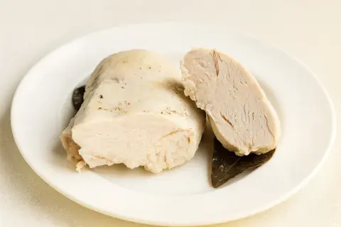 Un morceau de poulet poché dans un bouillon savoureux, coupé en deux pour montrer sa tendreté. Accompagné de feuilles de laurier.