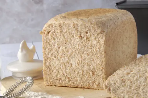 Un pain de farine intégrale, tout juste sorti du four, avec une tranche coupée. Des ingrédients de boulangerie sont disposés autour.