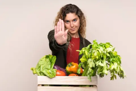 Femme faisant un geste d'arrêt devant une caisse de légumes frais, comme pour dire non aux légumes crus toxiques.
