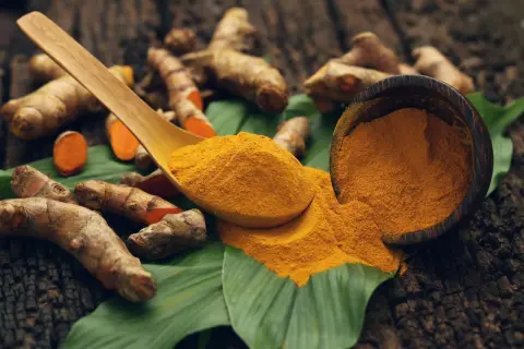 Racines de curcuma et poudre dorée sur une feuille verte, une cuillère en bois débordant de curcuma.