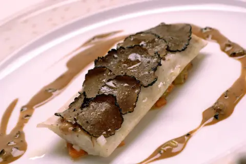 Un plat de poisson délicat, garni de fines lamelles de truffe de la Saint-Jean, arrosé d'une sauce onctueuse sur une assiette blanche.