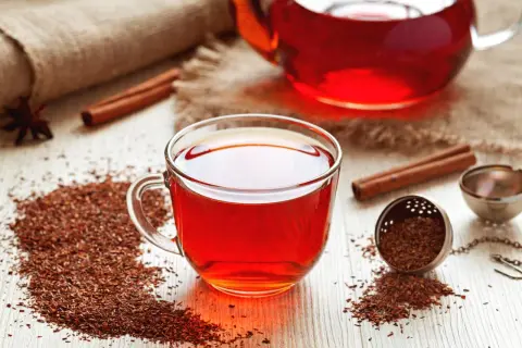 Une tasse de thé rooibos rouge vif, avec des bâtons de cannelle et des feuilles de thé sur une table en bois.