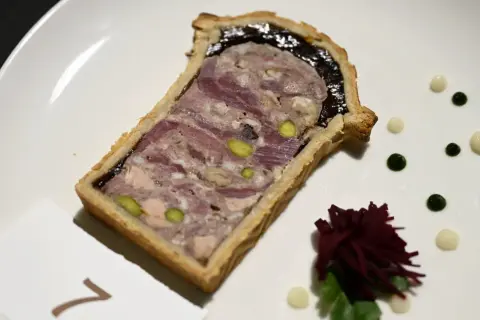Tranche de pâté en croûte, garnie de pistaches et d'une couche sombre, servie sur une assiette blanche avec des touches de sauce verte et blanche.