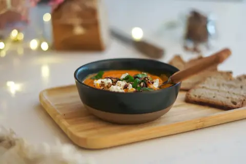 Un bol de soupe butternut tomate onctueuse, garnie de fromage frais et de noix, servi avec du pain grillé.