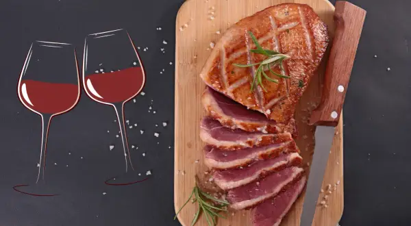 Un canard rôti tranché sur une planche en bois, accompagné de deux verres de vin rouge. La question se pose : quel vin avec le canard pour un repas parfait ?
