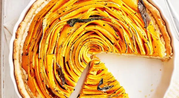 Une magnifique tarte à la courge aux fines lamelles dorées, garnie de feuilles de sauge. Une part a été retirée, révélant la texture appétissante.