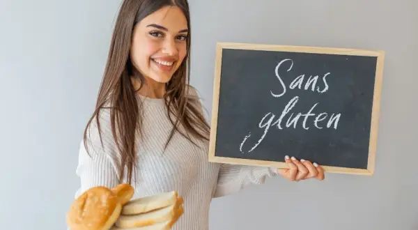 Une jeune femme souriante tient une assiette de pain et un tableau noir "Sans gluten". Elle montre ce qu'il faut manger en cas d'intolérance au gluten.