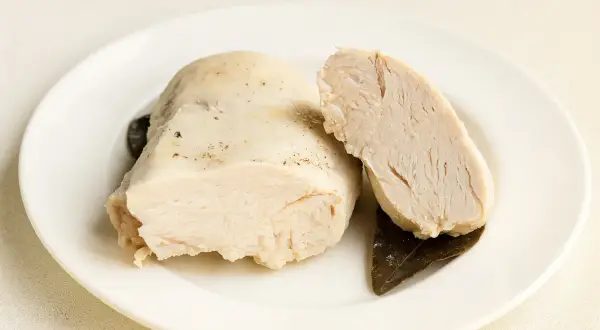 Un morceau de poulet poché dans un bouillon savoureux, coupé en deux pour montrer sa tendreté. Accompagné de feuilles de laurier.