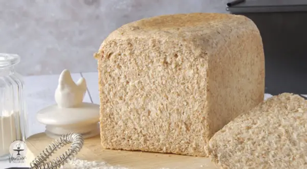 Un pain de farine intégrale, tout juste sorti du four, avec une tranche coupée. Des ingrédients de boulangerie sont disposés autour.