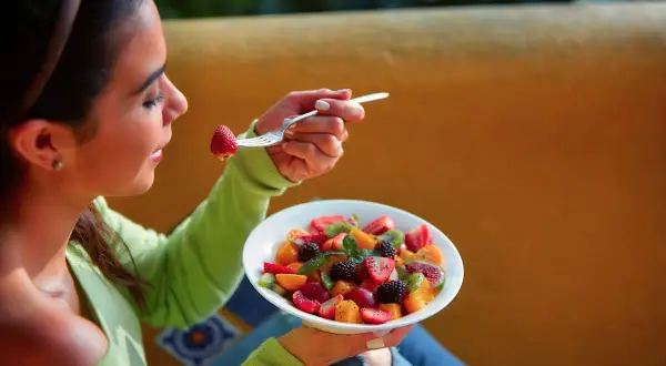 Une jeune femme savoure une salade de fruits colorée, une fraise à la fourchette. La naturopathie suggère quand manger les fruits pour une digestion optimale.
