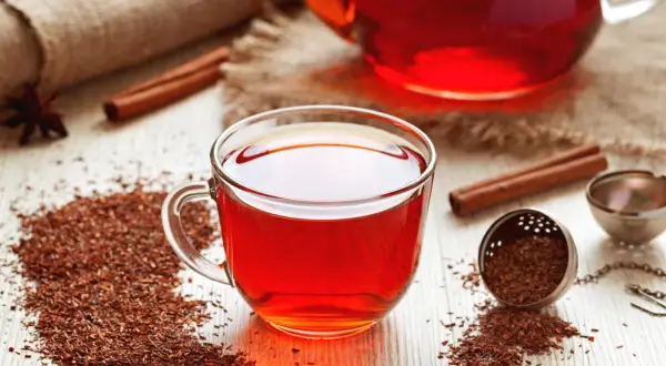 Une tasse de thé rooibos rouge vif, avec des bâtons de cannelle et des feuilles de thé sur une table en bois.