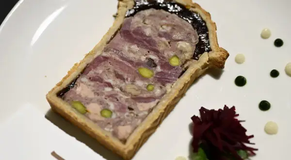 Tranche de pâté en croûte, garnie de pistaches et d'une couche sombre, servie sur une assiette blanche avec des touches de sauce verte et blanche.