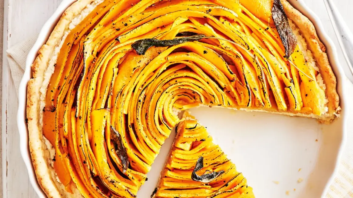 Une magnifique tarte à la courge aux fines lamelles dorées, garnie de feuilles de sauge. Une part a été retirée, révélant la texture appétissante.
