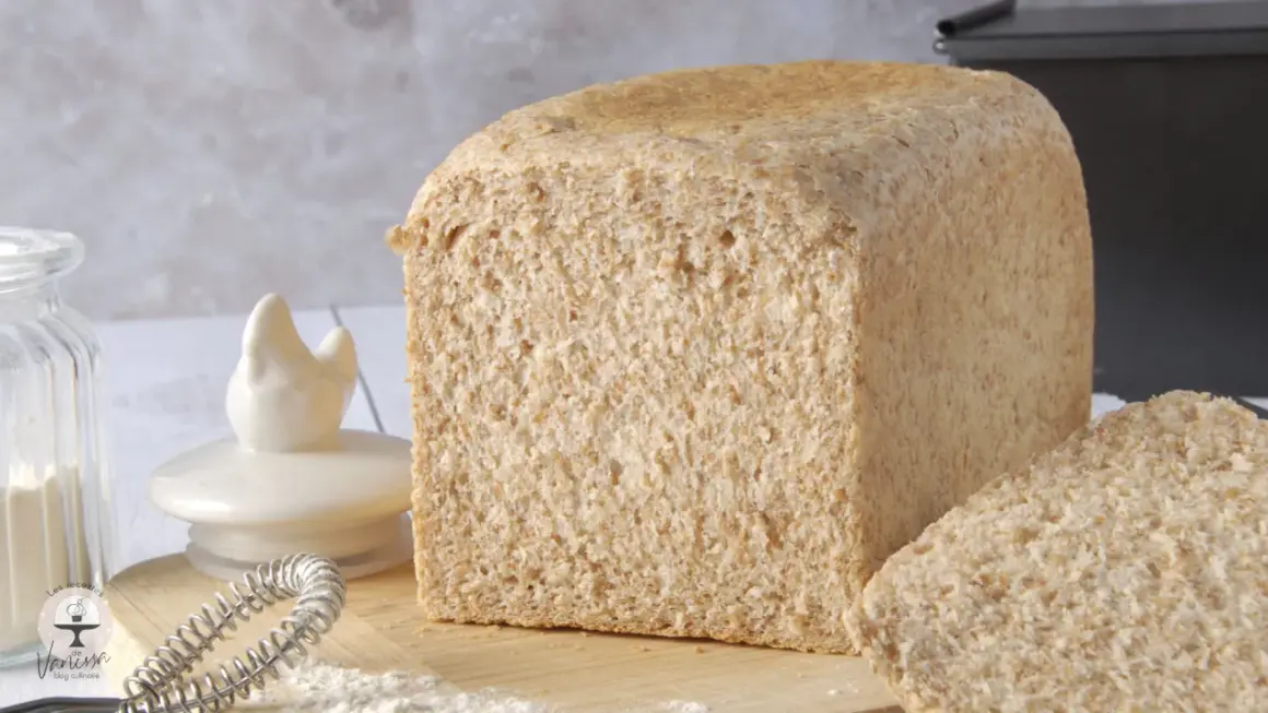Un pain de farine intégrale, tout juste sorti du four, avec une tranche coupée. Des ingrédients de boulangerie sont disposés autour.