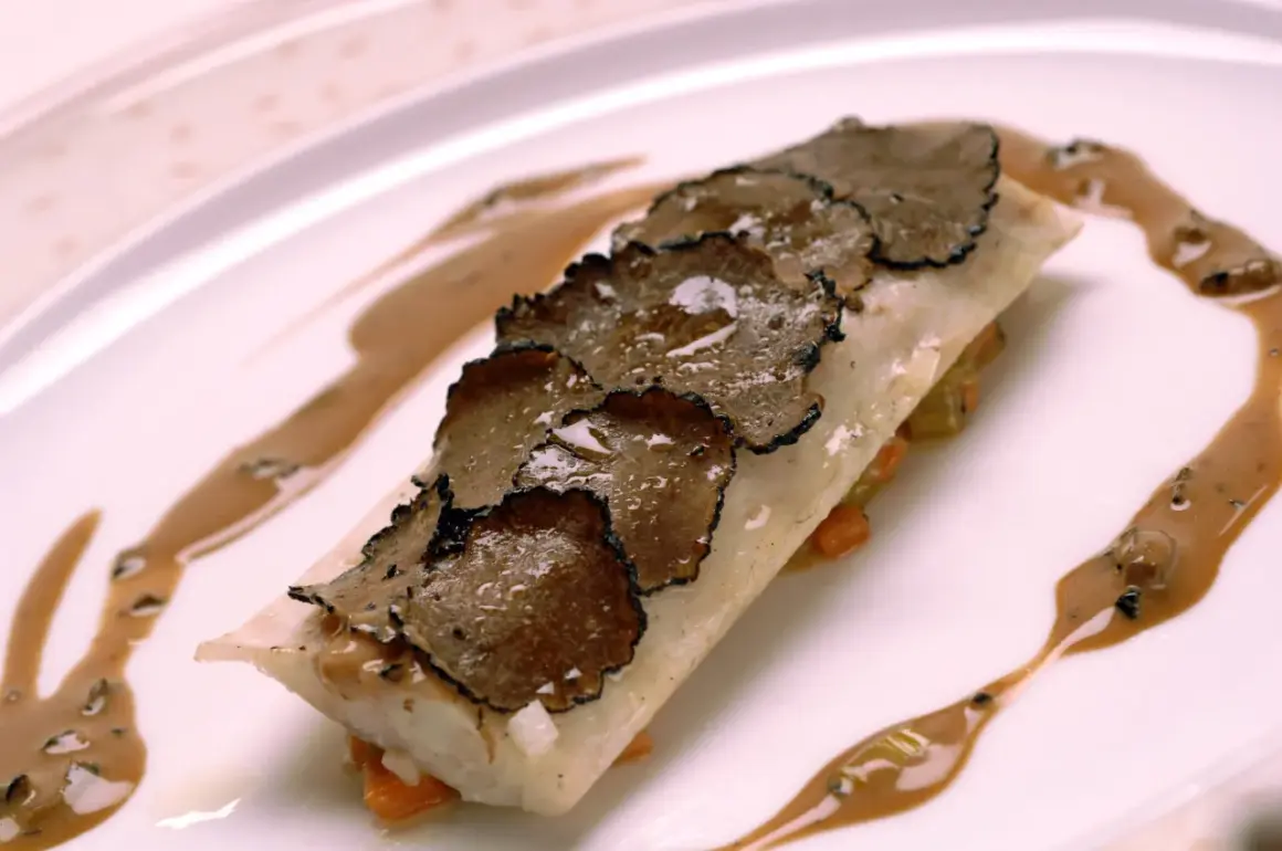 Un plat de poisson délicat, garni de fines lamelles de truffe de la Saint-Jean, arrosé d'une sauce onctueuse sur une assiette blanche.