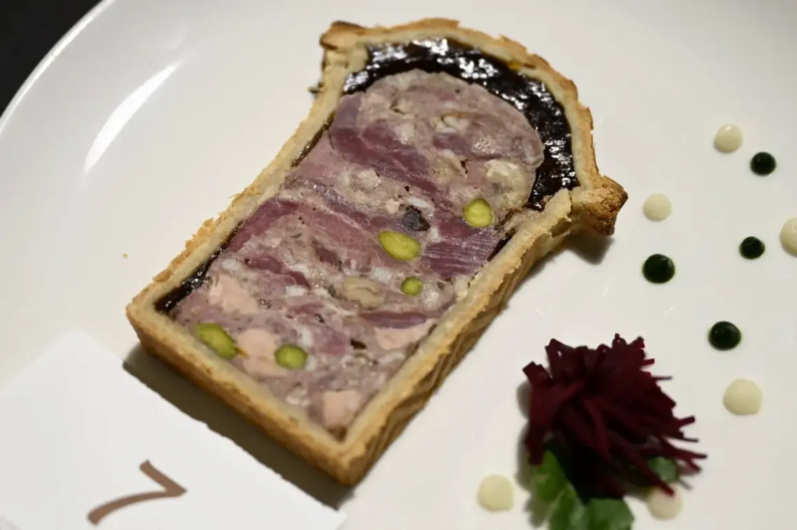 Tranche de pâté en croûte, garnie de pistaches et d'une couche sombre, servie sur une assiette blanche avec des touches de sauce verte et blanche.