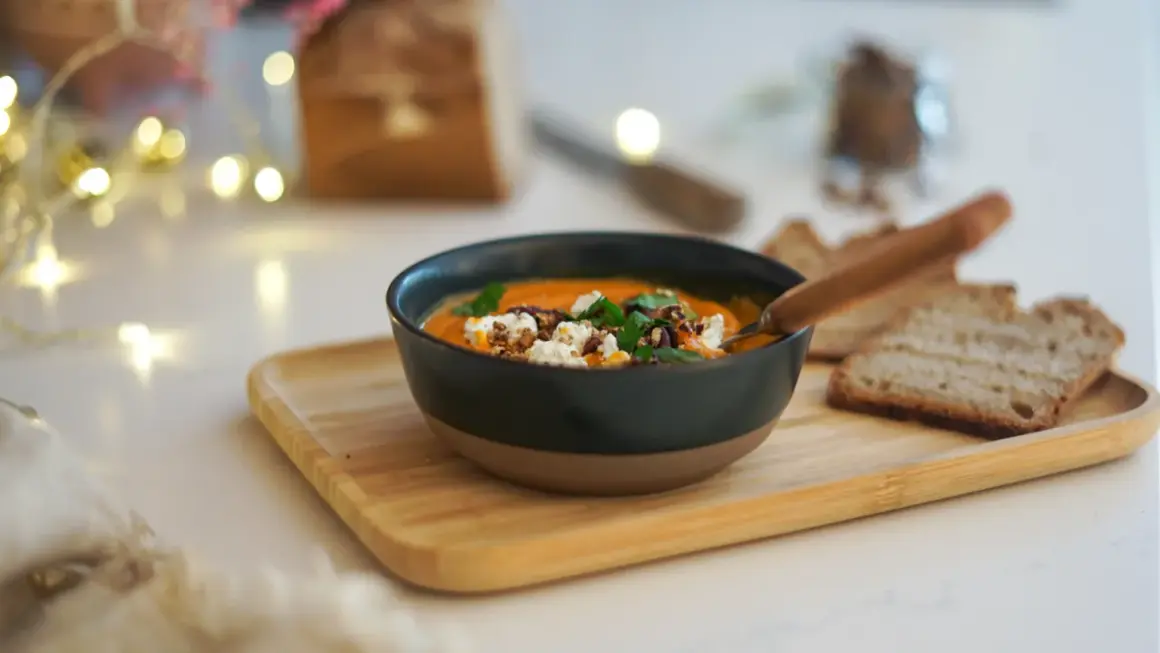 Un bol de soupe butternut tomate onctueuse, garnie de fromage frais et de noix, servi avec du pain grillé.