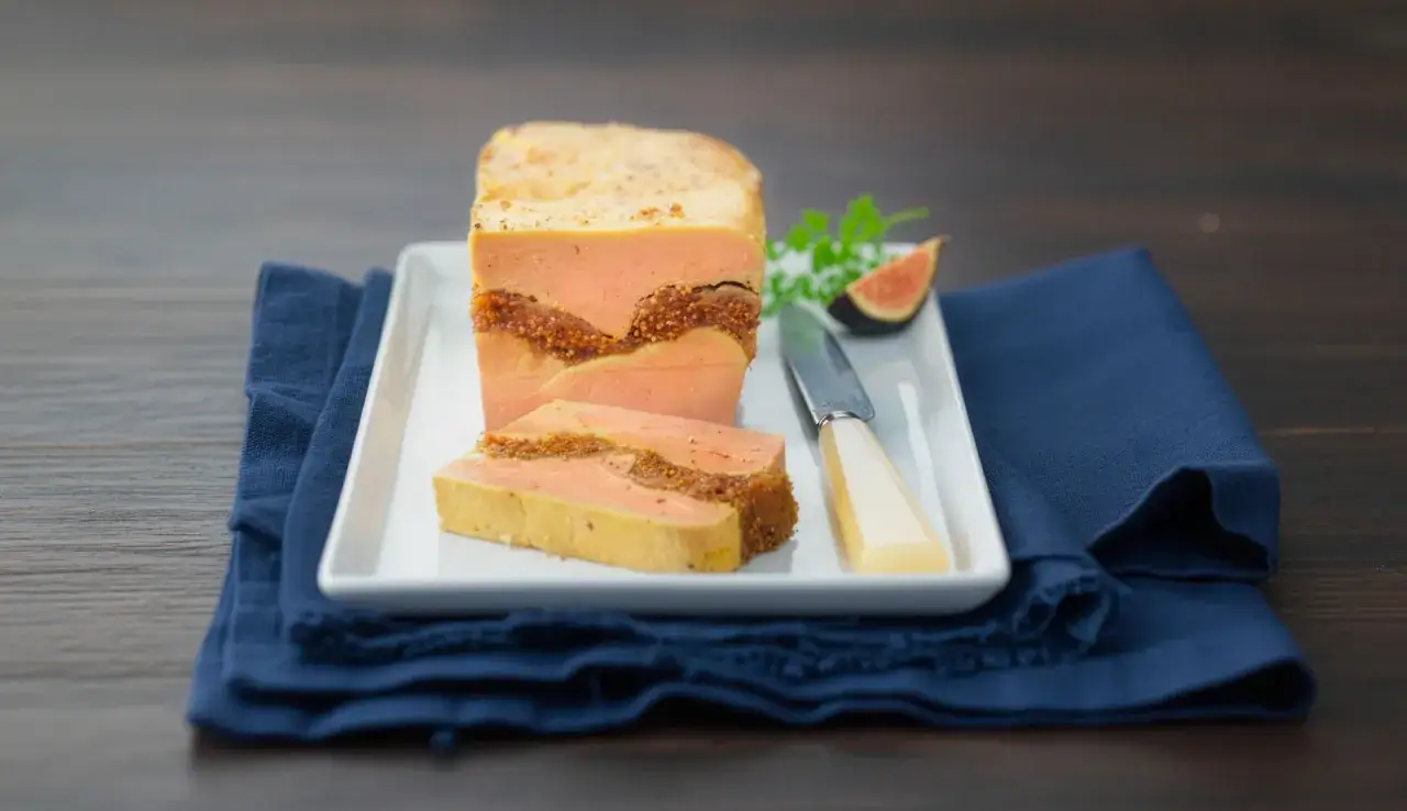 Tranche de figue au foie gras, une délicatesse qui se déguste aussi bien chaude que froide, présentée sur une assiette blanche avec une figue et un couteau.