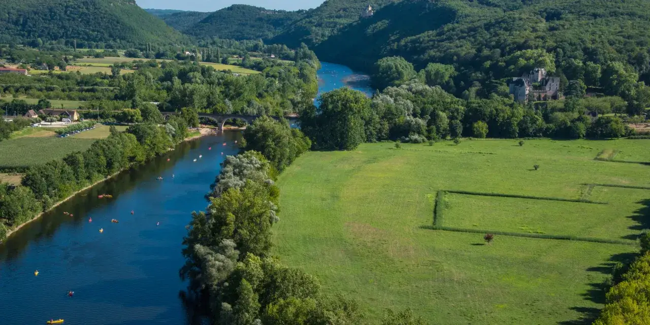 Paysage de la Dordogne, contrastant le vert des prairies et des for&ecirc;ts avec le bleu de la rivi&egrave;re o&ugrave; naviguent des kayaks. Le ch&acirc;teau de Castelnaud se dresse au loin.