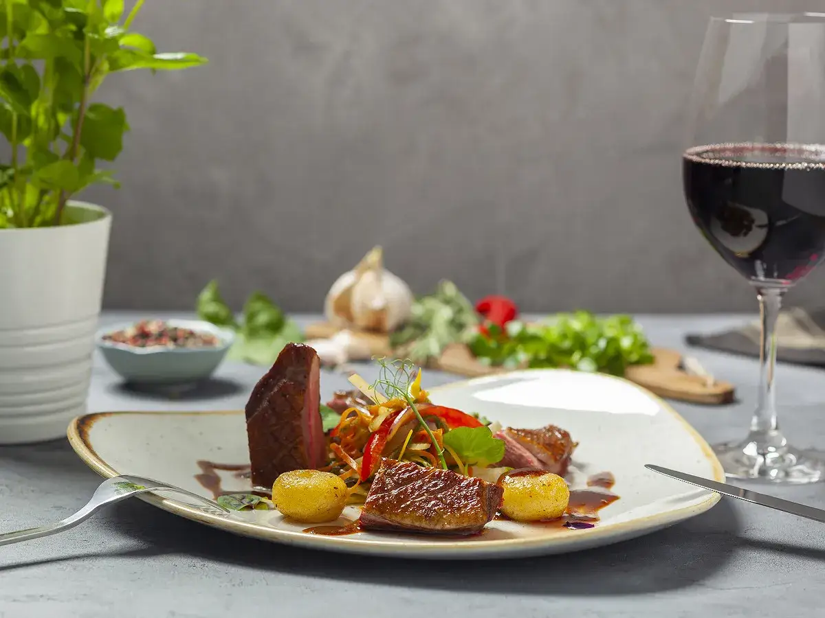 Un plat de canard savoureux avec des pommes de terre et des légumes, accompagné d'un verre de vin rouge. La question est : quel vin avec le canard ?