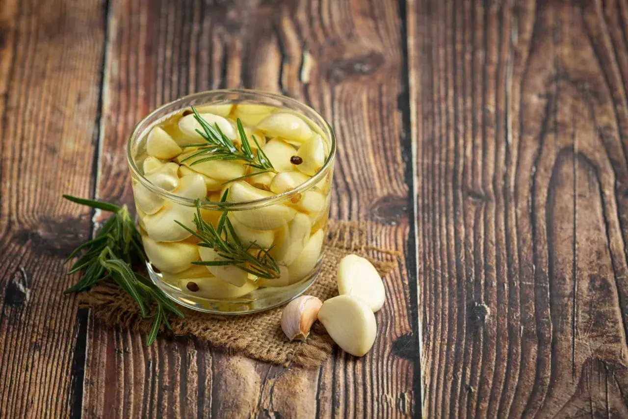Un bocal de gousses d'ail confit avec du romarin et des grains de poivre sur une table en bois. Une id&eacute;e pour faire de l'ail confit.