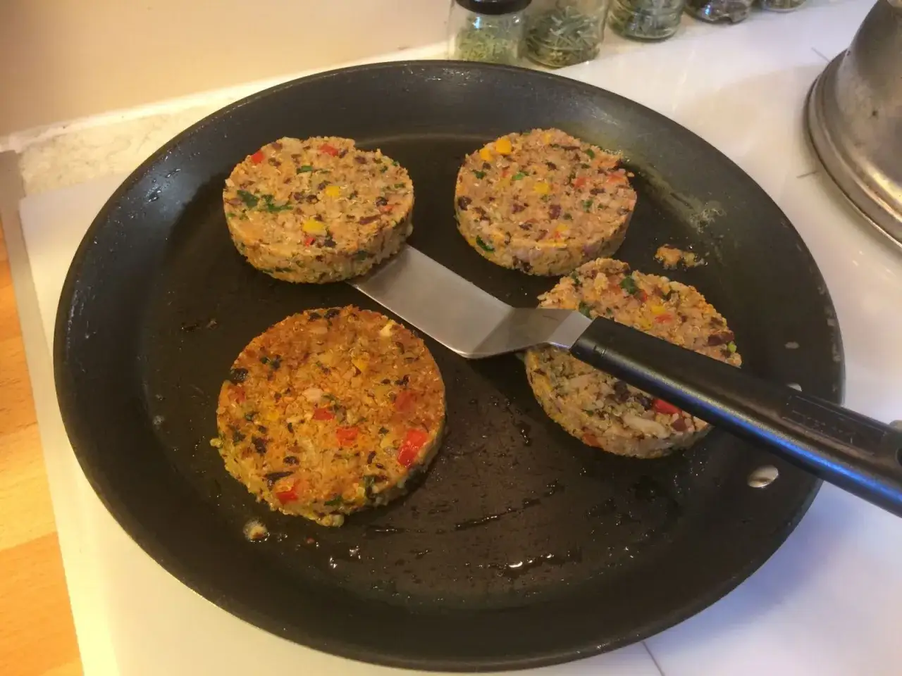 Quatre galettes de haricot rouge, colorées et garnies de légumes, dorent dans une poêle avec une spatule.