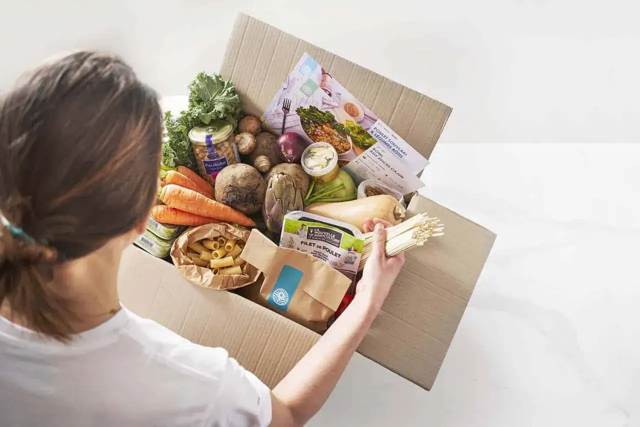 Une personne déballe une boîte remplie d'ingrédients frais : carottes, betteraves, courge, poulet, pâtes et pois chiches. Idéal pour un budget courses pour 2 personnes par semaine.