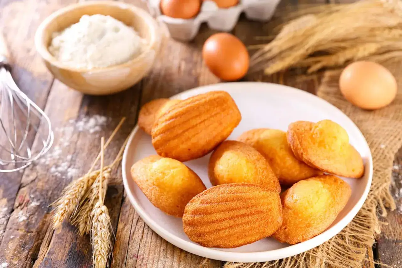 Une assiette de madeleines dor&eacute;es, &eacute;voquant la madeleine de Proust recette, avec des ingr&eacute;dients comme la farine, les &oelig;ufs et du bl&eacute;.
