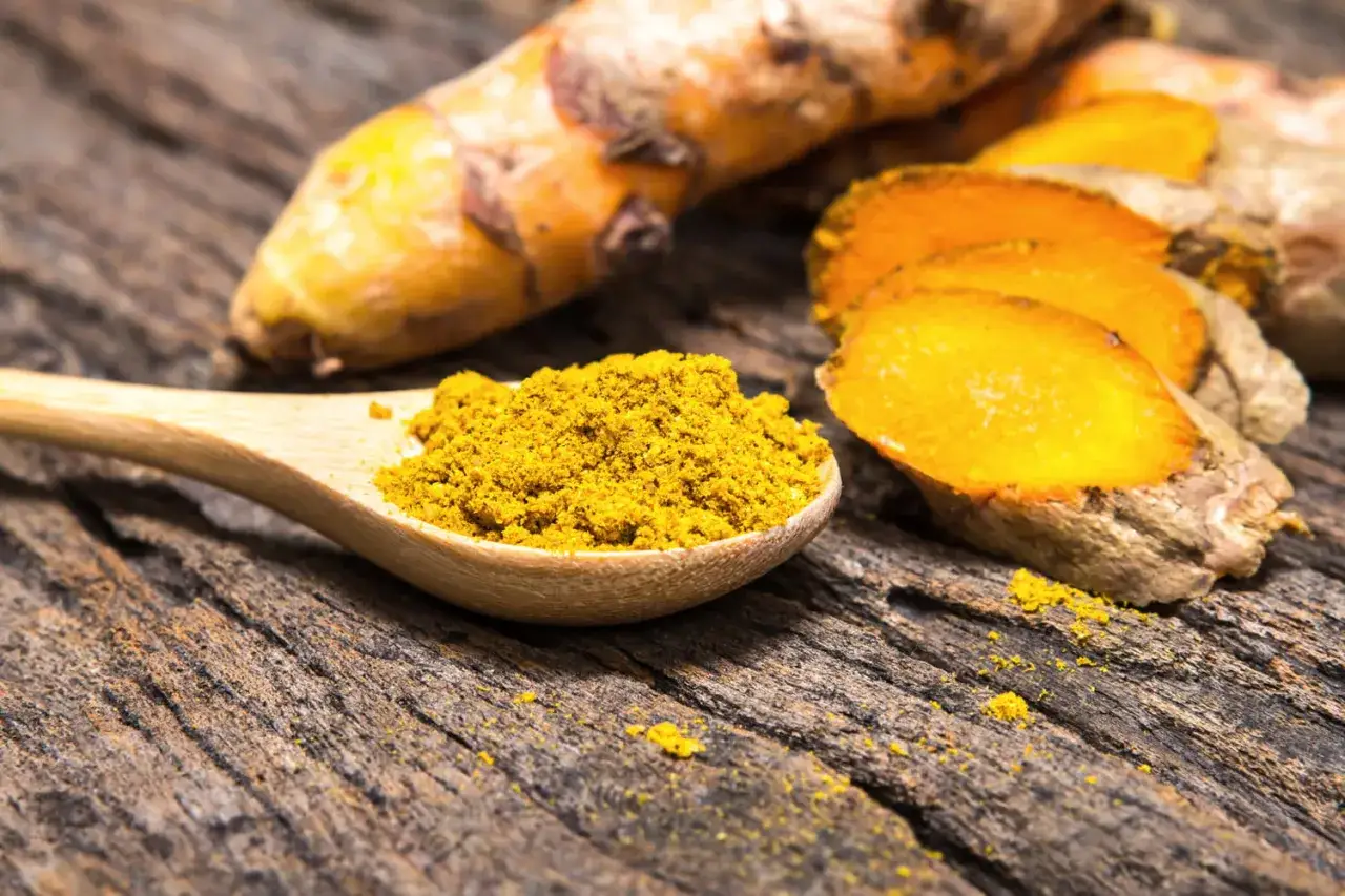 Une cuill&egrave;re en bois d&eacute;borde de poudre de curcuma, &agrave; c&ocirc;t&eacute; de racines coup&eacute;es en tranches sur un fond de bois vieilli.