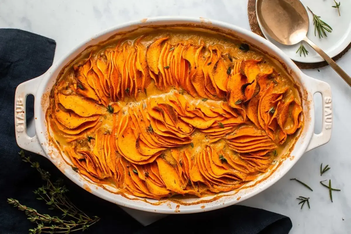 Gratin de patate douce, une recette sans gluten, aux fines tranches orange vif, baignant dans une sauce cr&eacute;meuse et garnie d'herbes fra&icirc;ches.