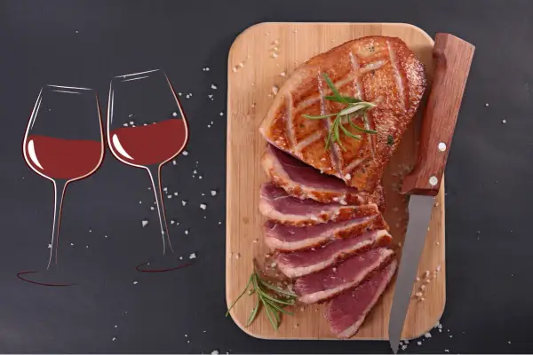 Un canard rôti tranché sur une planche en bois, accompagné de deux verres de vin rouge. La question se pose : quel vin avec le canard pour un repas parfait ?