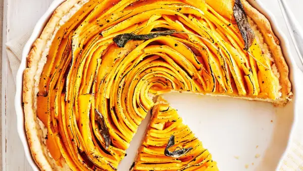 Une magnifique tarte à la courge aux fines lamelles dorées, garnie de feuilles de sauge. Une part a été retirée, révélant la texture appétissante.